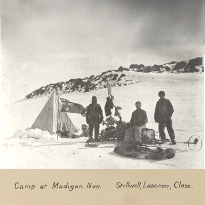 Black and white photo of three men wearing cold-weather clothing in a very snowing environment with their equipment, a tent and an Australian flag.